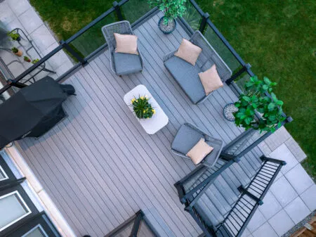 Aerial view of a Fiberon Chai deck with Fortress-Railings1 showcases gray flooring. Features include a seating area with gray chairs and beige cushions, a white coffee table with plants, another decorative plant, and a covered grill. Its all beautifully enclosed by a wooden fence and lush lawn.