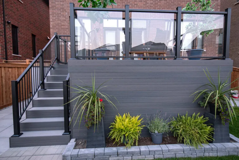 A modern Fiberon Chai deck features gray flooring and a seating area with four gray chairs, each adorned with a beige cushion. A white rectangular table holds a potted plant with yellow flowers. The deck, encompassed by Fortress Railings4 and a wooden fence, offers contemporary charm.