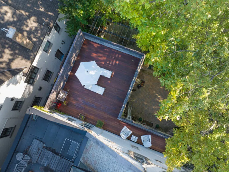 Aerial view of an IPE deck featuring a white patio umbrella and reclining chairs, complemented by additional seating along the perimeter. The area is framed by a gray railing, with lush green trees providing a serene backdrop.