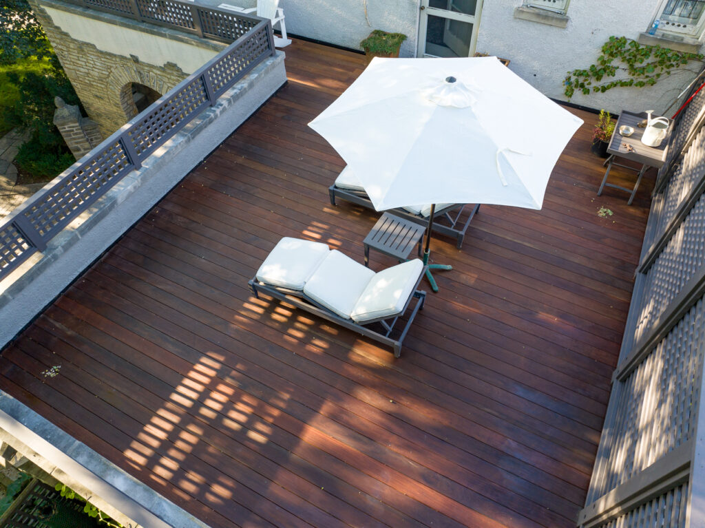 Aerial view of an IPE deck featuring a white patio umbrella and reclining chairs, complemented by additional seating along the perimeter. The area is framed by a gray railing, with lush green trees providing a serene backdrop.