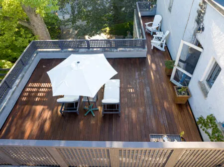 Aerial view of an IPE deck featuring a white patio umbrella and reclining chairs, complemented by additional seating along the perimeter. The area is framed by a gray railing, with lush green trees providing a serene backdrop.