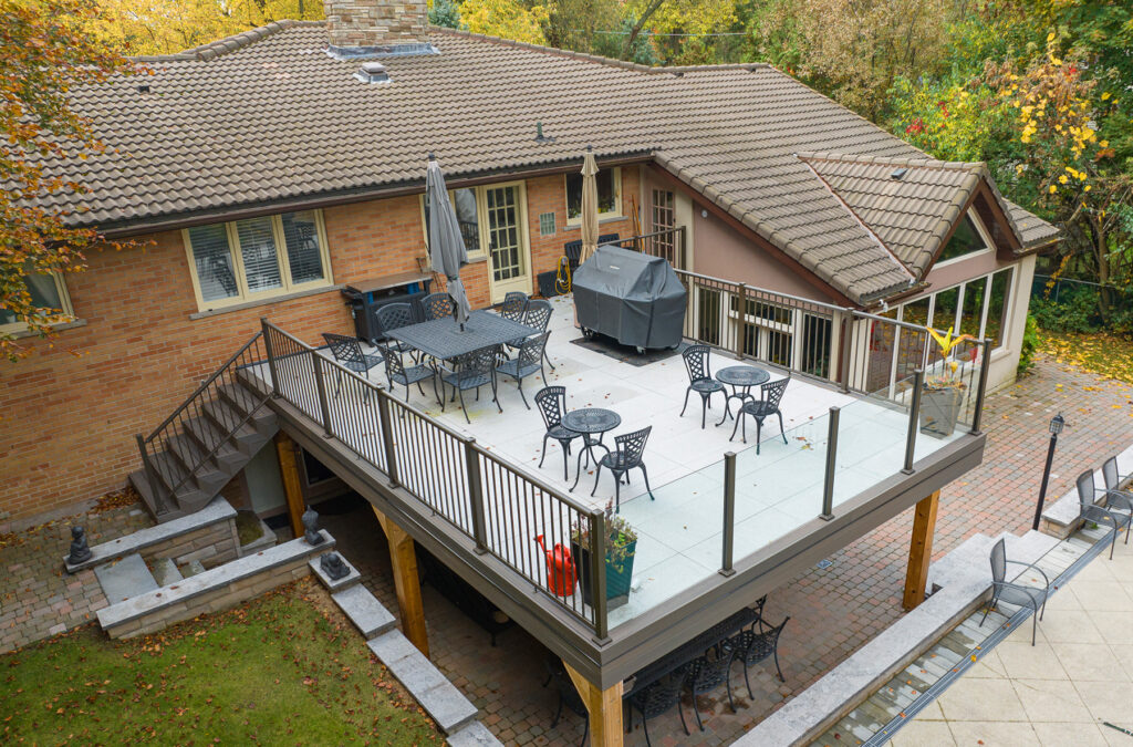 Aerial view of a suburban home showcases a spacious elevated deck featuring professionally installed metal railings by experts in Toronto. The area includes a grill, three small tables with chairs, and a larger dining table. Surrounding trees display vibrant autumn foliage as steps lead to the brick patio below.