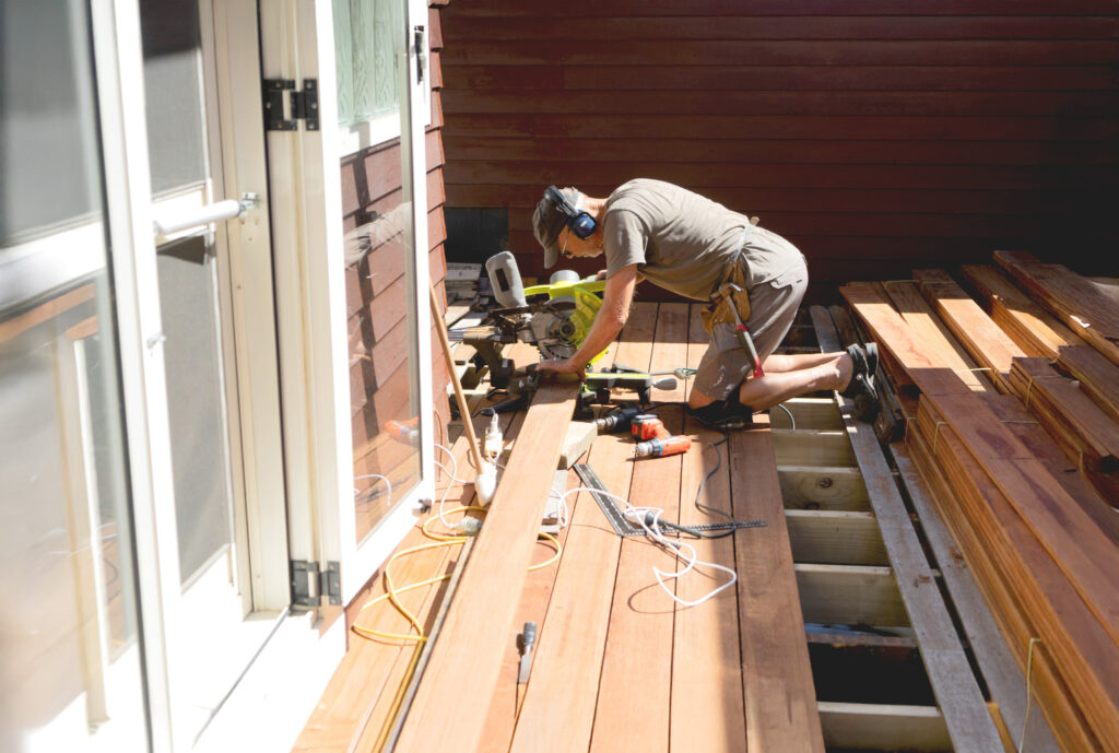 A person wearing shorts, a t-shirt, and headphones is working with a saw on a wooden deck. Various tools and wooden planks are scattered around. The area is brightly lit, suggesting daytime.