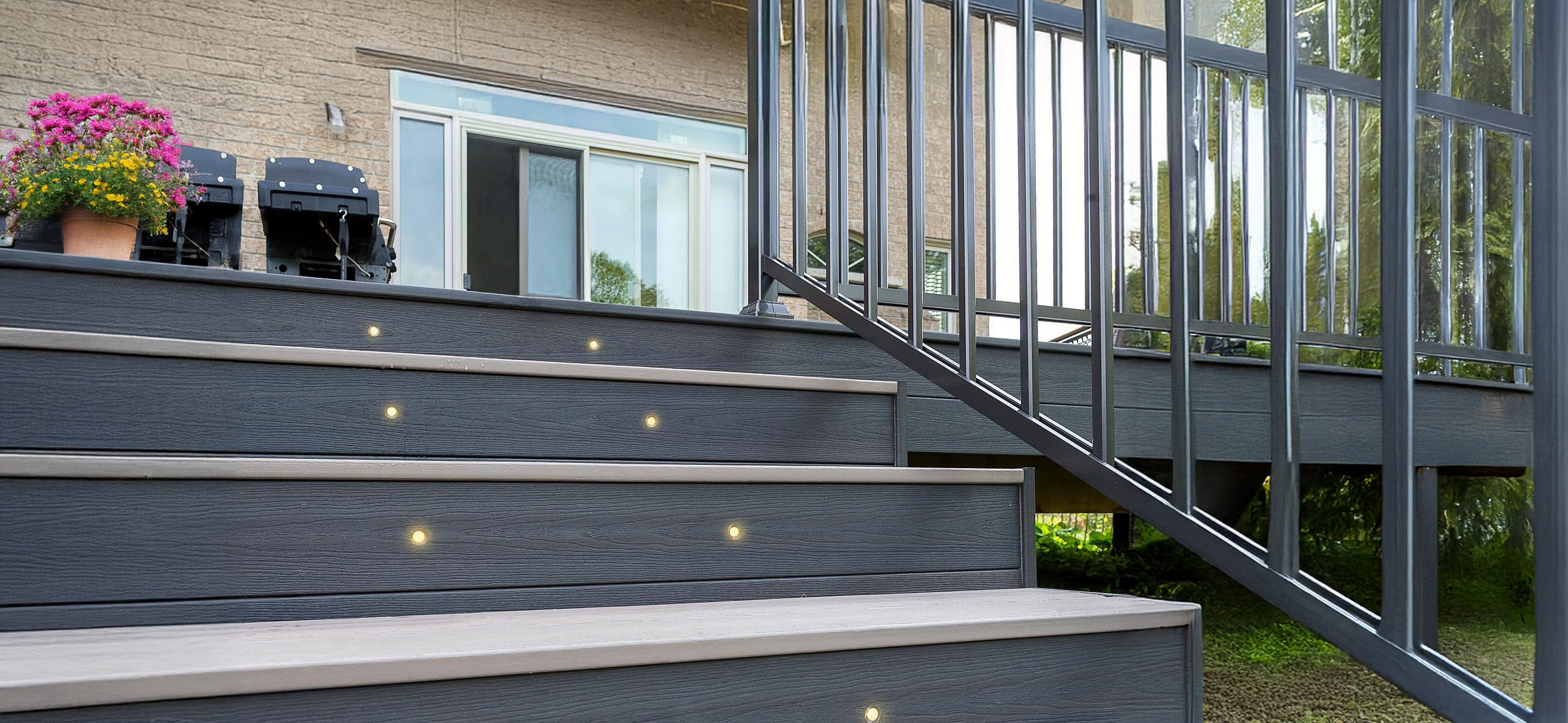 Outdoor stairs, adorned with built-in lights, lead up to a deck, where sleek metal railings flank the steps. A potted plant with pink flowers sits near a glass sliding door that opens to the house, while a barbecue grill awaits on the well-lit deck.