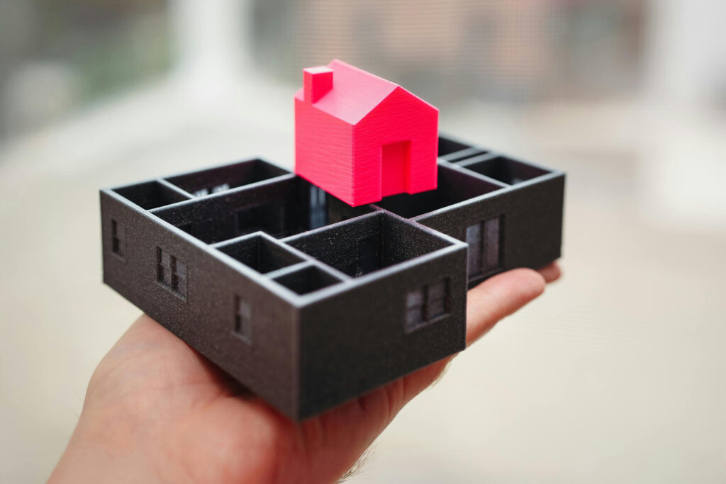 A hand holding a small architectural model showing a black, open-layout building with dividers, and a red, simplified house structure on top. The background is blurred, highlighting the models details.