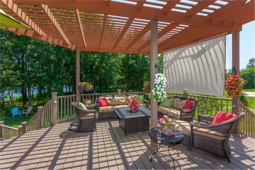 A wooden patio deck features a pergola casting striped shadows. It has wicker chairs and a sofa with floral cushions surrounding a central fire pit. Flower pots hang from the pergola and are placed on a small side table. Trees are visible in the background.