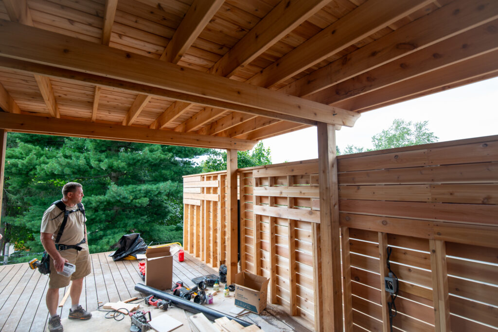 A rooftop deck builder in Toronto stands on a wooden deck under construction, surrounded by various tools and materials. The structure features a roof with partially completed walls, set against a backdrop of lush green trees.