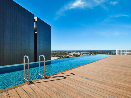 A rooftop infinity pool with clear blue water and ladder sits gracefully on composite decking, encircled by sleek wooden tones. The modern black building structure stands against a clear blue sky, offering a stunning backdrop of the distant cityscape.