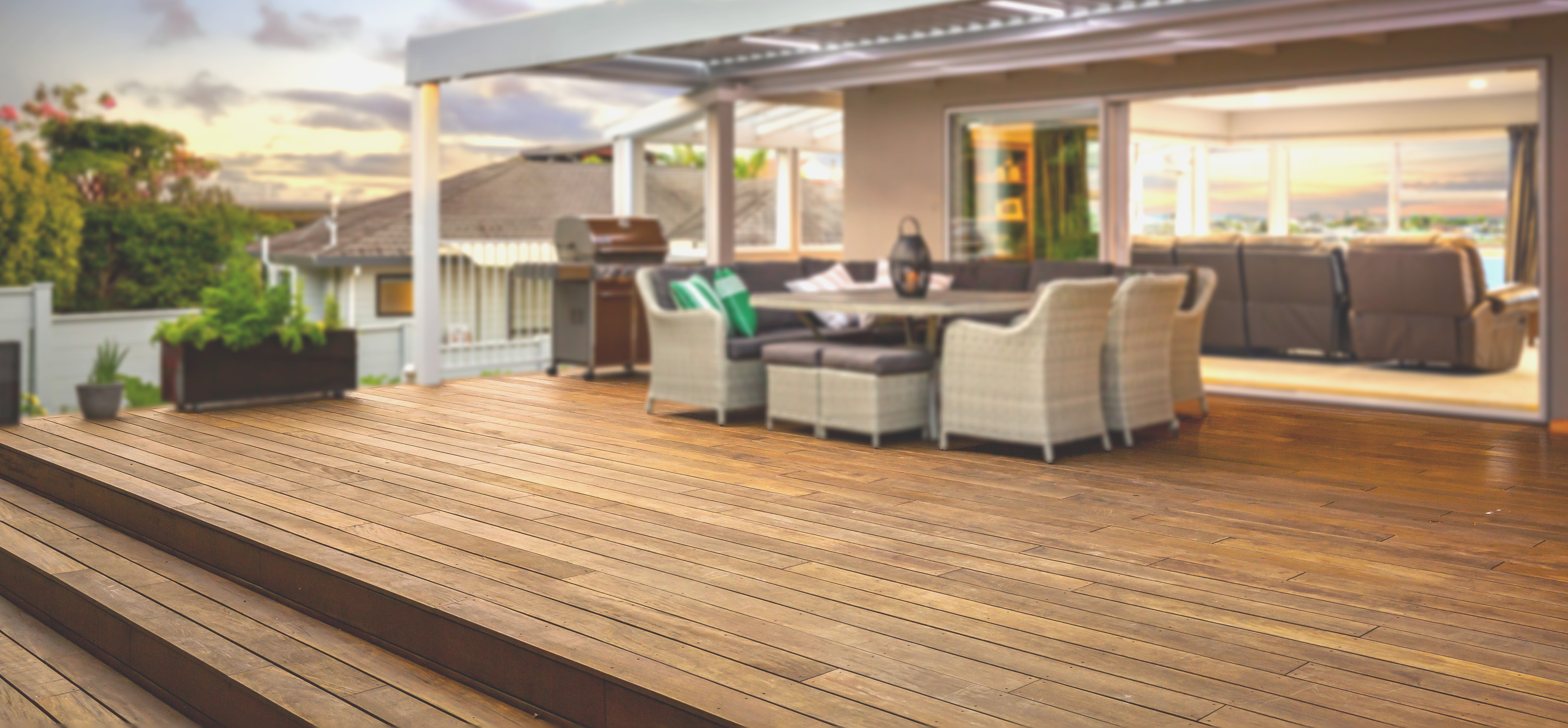 Spacious wooden deck crafted by a renowned Deck Builder Toronto, featuring an outdoor dining set under a pergola, adjacent to a contemporary home. Open sliding doors reveal a cozy interior with sofas. Lush greenery surrounds the area as the sky is partly cloudy at sunset.