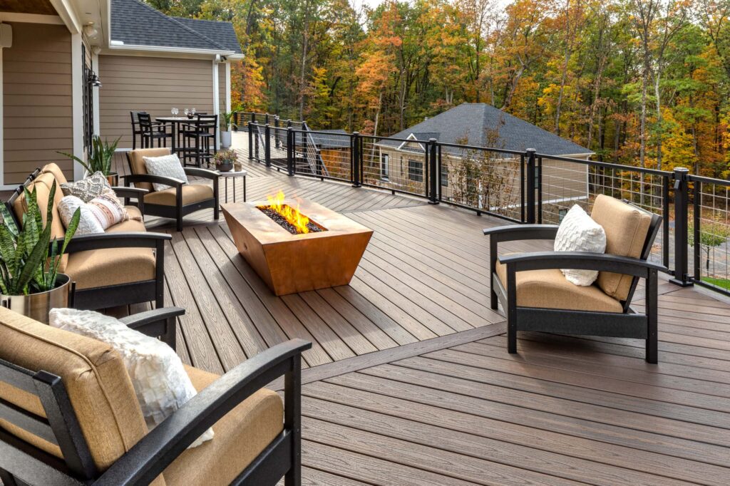 Spacious wooden deck featuring a modern fire pit with a lively flame. Surrounded by cushioned chairs with throw pillows, the deck overlooks an autumn forest. A dining table with chairs is visible in the background near the house.
