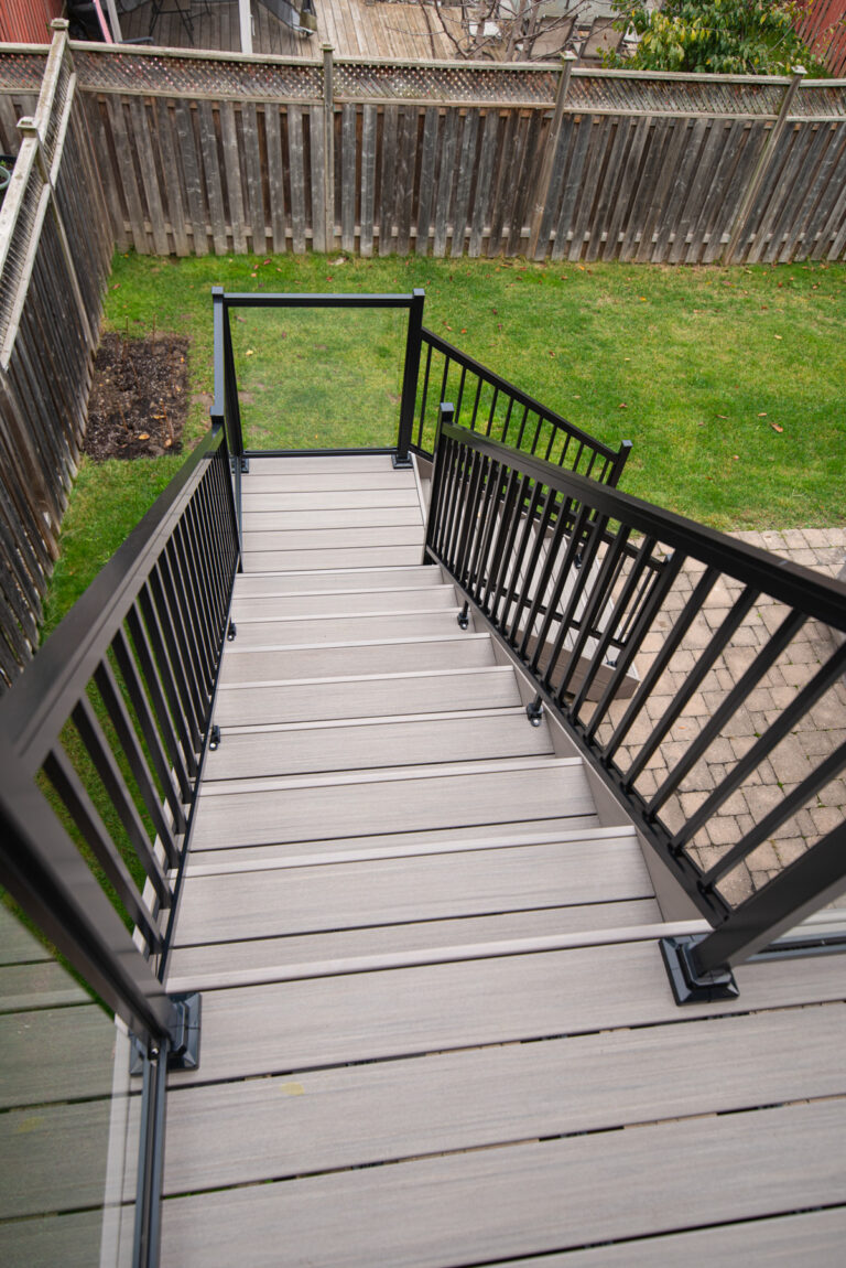 A view looking down a set of modern outdoor stairs with gray composite decking and black metal railings, leading from a deck to a grassy backyard enclosed by a wooden fence.