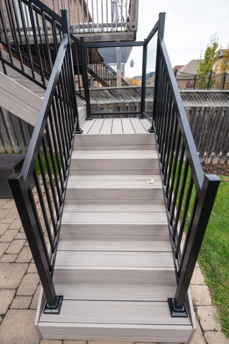 Gray outdoor stairs with black metal railings leading up to a deck. The stairs are surrounded by grass and paving stones, and a wooden fence is visible in the background.