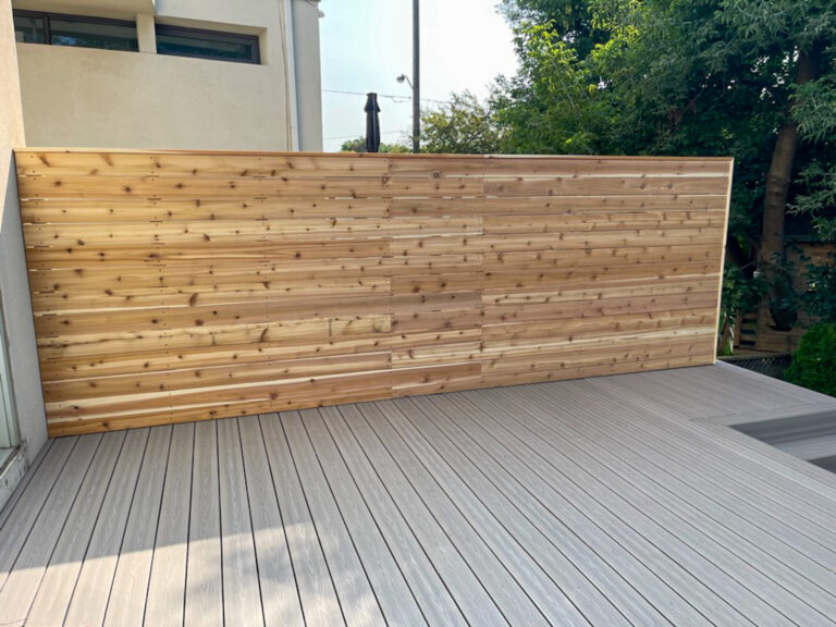 A newly constructed cedar privacy screen stands on light-colored composite decking in a modern outdoor space, with trees and a building visible in the background.