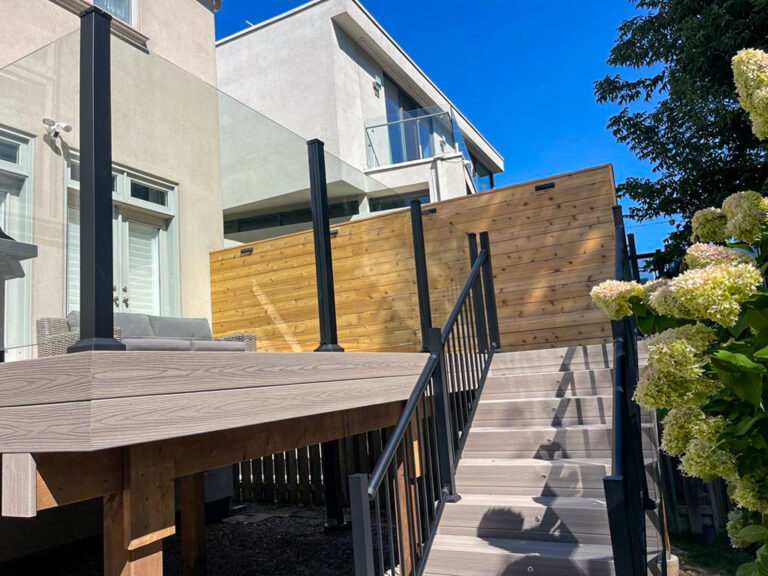 Modern backyard deck with glass railing, wooden privacy wall, and stairs leading up. Bright sunlight, blue sky, and blooming white hydrangeas are visible in the foreground.