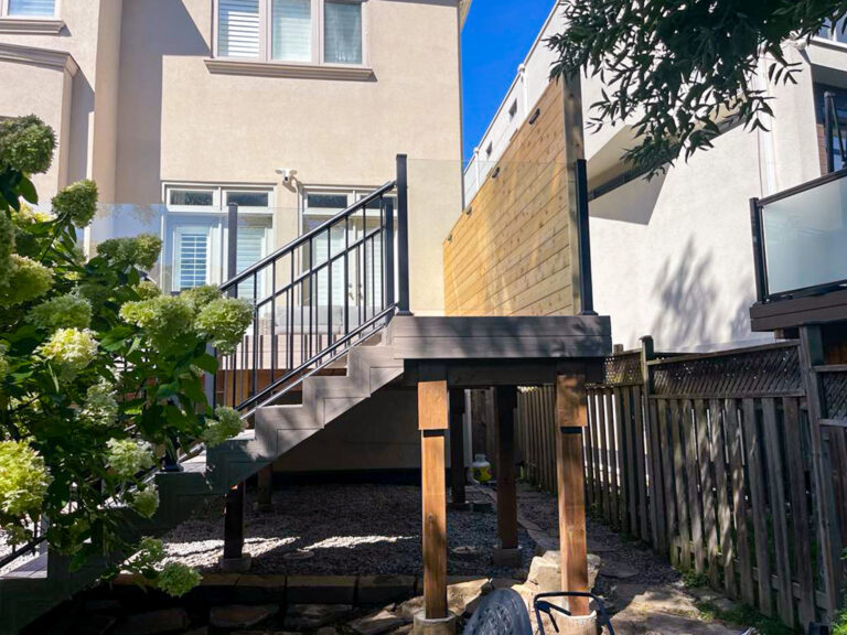 A modern raised deck built with Fiberon Composite Decking features black railings and stairs, attached to the back of a beige house and overlooking a small gravel yard with plants, trees, and a wooden fence.
