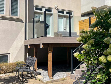 A modern backyard patio with glass railings, a small outdoor sofa, and wooden support beams. A metal chair and flowers are in the foreground, and stairs lead up to the patio. Sunlight casts shadows across the area.
