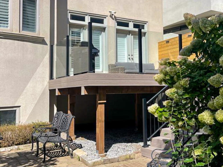 A modern backyard patio with glass railings, a small outdoor sofa, and wooden support beams. A metal chair and flowers are in the foreground, and stairs lead up to the patio. Sunlight casts shadows across the area.