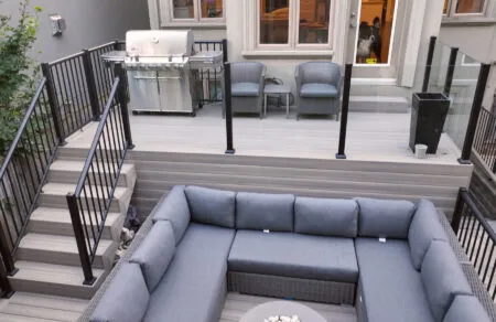 A modern outdoor patio with Fiberon Composite Decking features a sunken seating area where gray cushioned wicker sofas form an L-shape around a round coffee table, surrounded by stairs and sleek black railings.