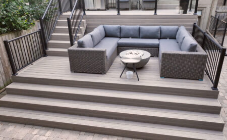 A modern outdoor patio with Fiberon Composite Decking features a sunken seating area where gray cushioned wicker sofas form an L-shape around a round coffee table, surrounded by stairs and sleek black railings.
