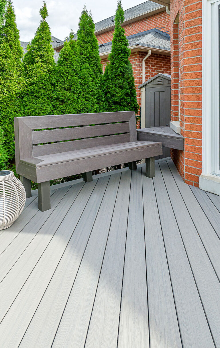 A modern gray bench with a backrest sits on a light gray composite deck near a red brick house, with tall green shrubs and a decorative white lantern beside it.