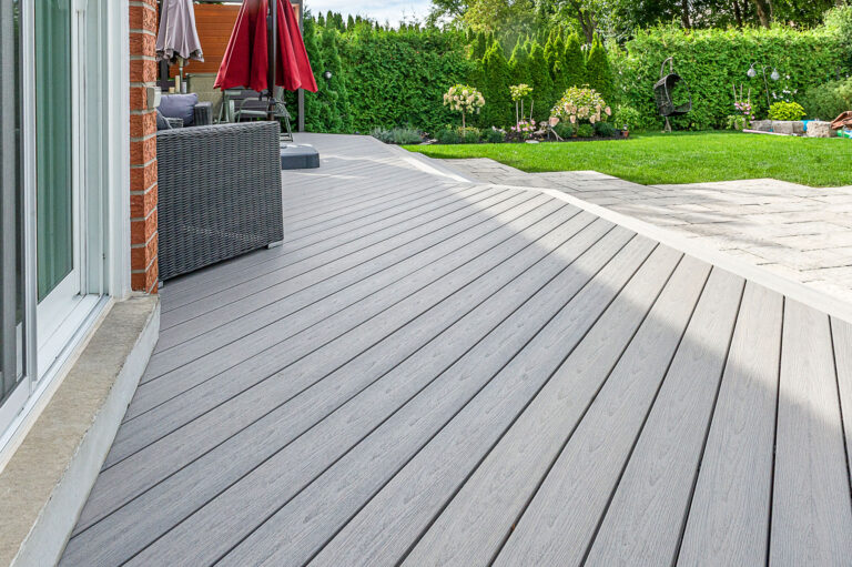 A modern backyard with a light gray composite deck beside a house, leading to a patio area with furniture and red umbrellas. The yard features green grass, stone paving, and lush hedges in the background.