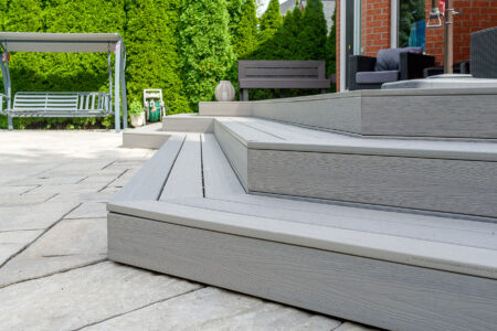 Close-up of wide, light gray composite deck steps leading from a patio to a seating area, with outdoor furniture and green hedges in the background.