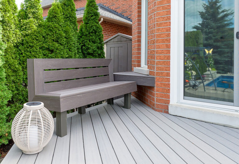 Gray wooden bench and white lantern on a light gray deck next to a red brick house, with tall green shrubs and a glass door reflecting an outdoor pool area.
