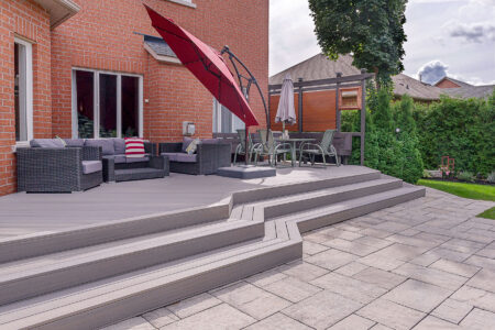 A backyard patio featuring gray composite decking, outdoor sofa set, red umbrella, dining table with umbrella, and stone paving. A brick house and lush greenery create a charming backdrop.
