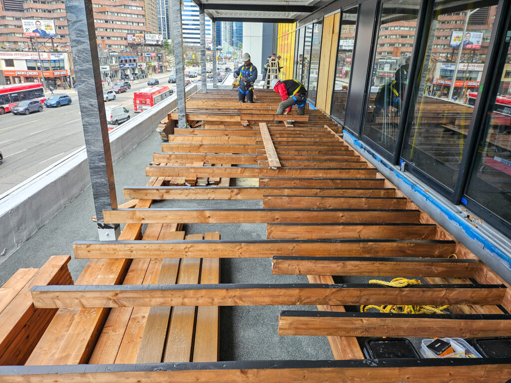 Delta Decks at work: Construction workers are building a wooden deck on a city balcony near the Cactus Club Cafe patio. Planks and tools are scattered around, with tall buildings and a busy street visible in the background.