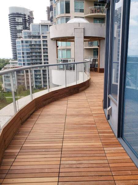 Curved IPE wood balcony with glass railing on a high-rise, overlooking modern city buildings in the background.