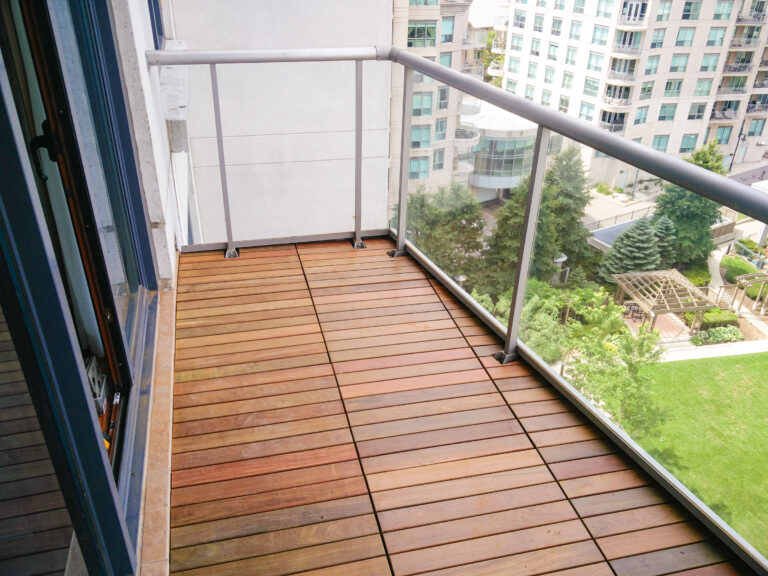 Wooden balcony with glass railing overlooks a courtyard with green trees and modern apartment buildings in the background.