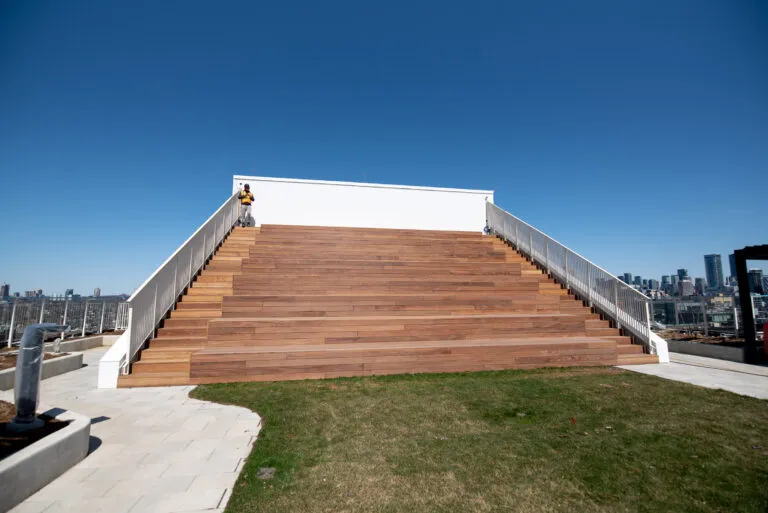 Large wooden steps lead up to a rooftop platform with metal railings; a person stands at the top under a clear blue sky.