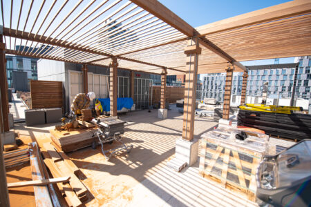 A construction worker cuts wood under a pergola structure on a rooftop surrounded by building materials.