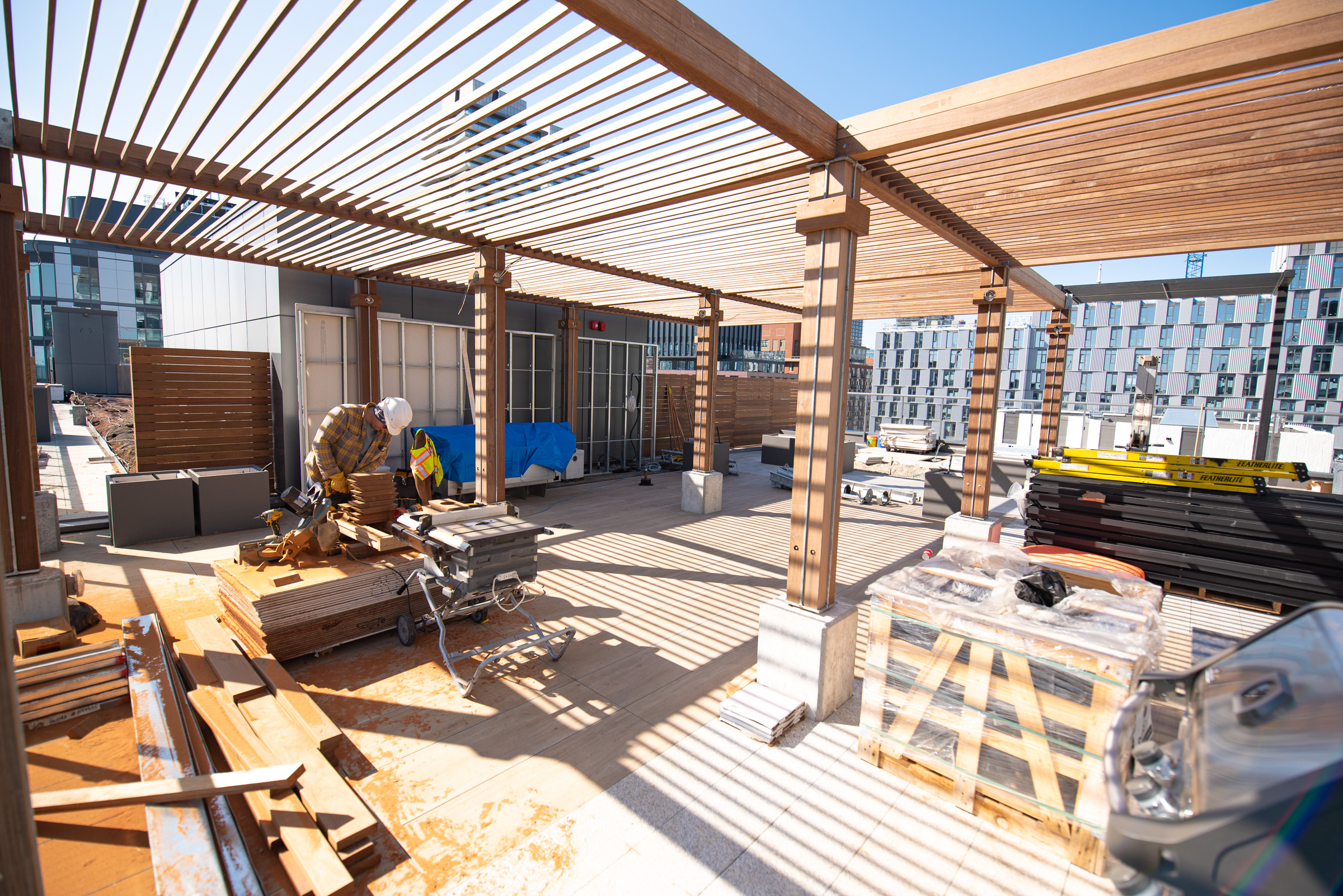 A construction worker cuts wood under a pergola structure on a rooftop surrounded by building materials.