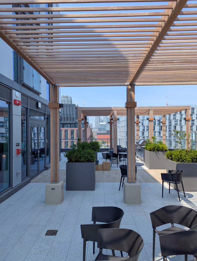 Rooftop patio with wooden pergola, black chairs, tables, and planters, overlooking a cityscape in the background.