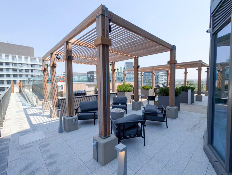 Modern rooftop terrace with pergolas, seating areas, planters, and city buildings in the background on a clear day.