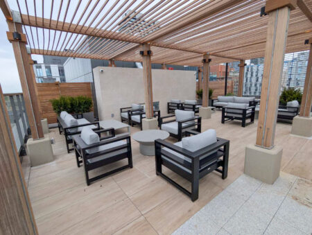 Rooftop patio with wooden pergola, gray cushioned chairs, sofas, and round tables, overlooking modern buildings.