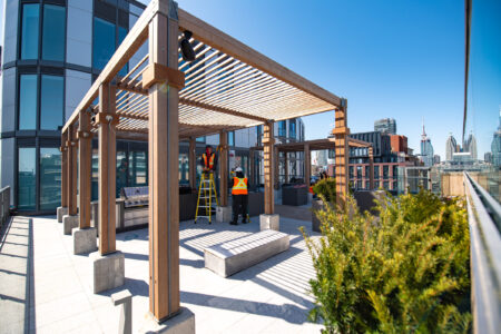 Workers in safety vests stand under a wooden pergola on a rooftop, with city buildings visible in the background.