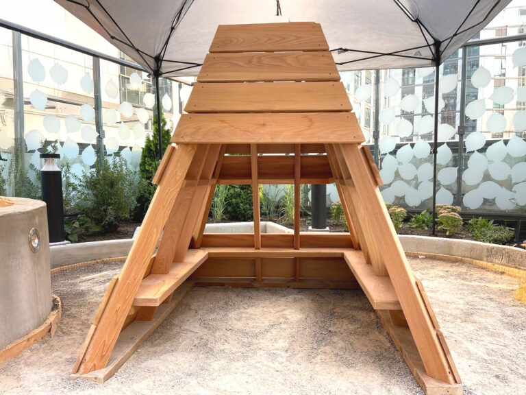 Wooden A-frame structure with benches inside, set outdoors under a canopy on a concrete surface.