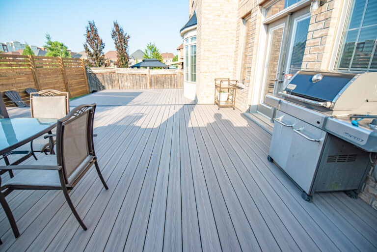 Spacious backyard patio with gray composite decking, outdoor dining set, grill, and wooden privacy fence on a sunny day.