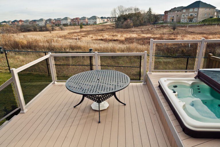 A round metal patio table and a hot tub on a deck overlook a field with houses in the distance.