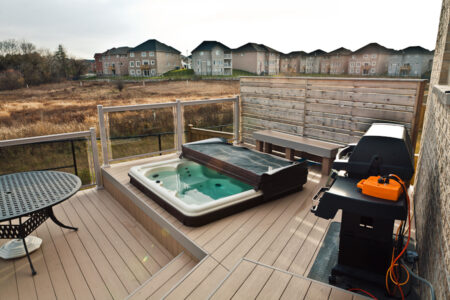 A backyard deck with a hot tub, barbecue grill, round table, bench, and fenced view of residential buildings and open field.