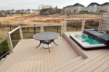 A backyard deck with a round metal table, built-in hot tub, glass railing, and a view of a grassy field and houses.
