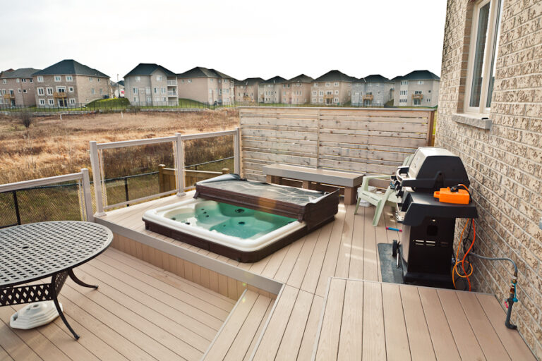 A backyard deck with a hot tub, grill, table, bench, and wooden privacy screen overlooking a suburban neighborhood.