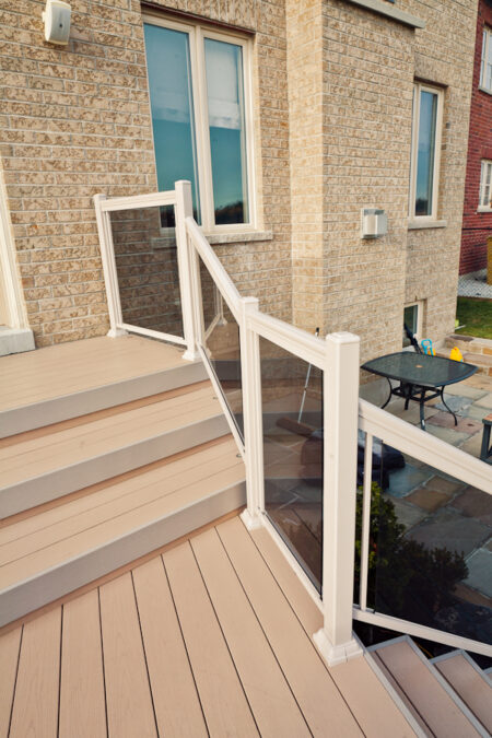 Light-colored deck stairs with white railings and glass panels, attached to a brick house, overlooking a patio area.