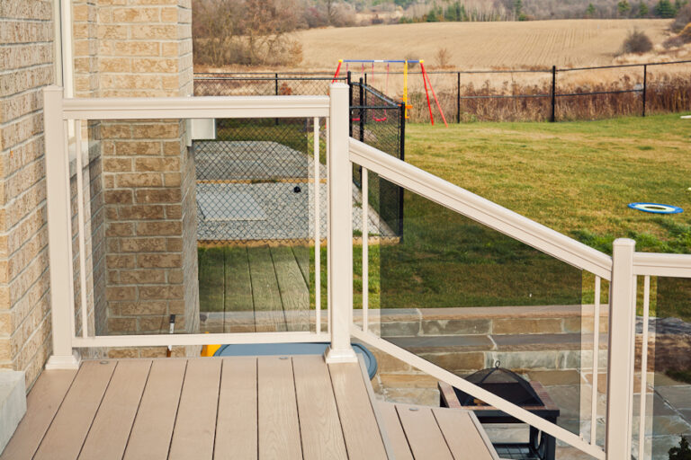 View from a deck with glass railing, overlooking a backyard with a swing set and open field in the background.