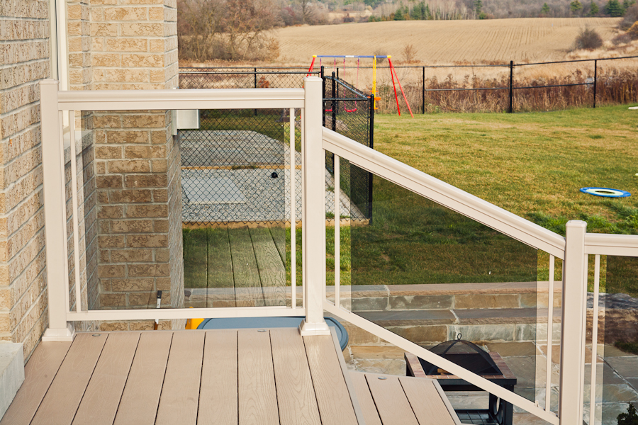 View from a deck with glass railing, overlooking a backyard with a swing set and open field in the background.