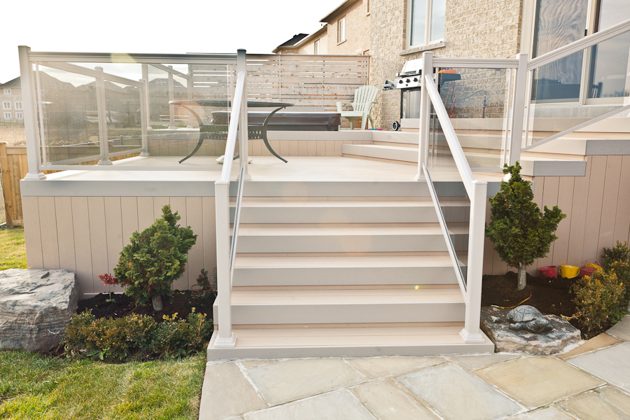 Backyard deck with glass railings, stairs, patio furniture, small trees, and stone landscaping in a residential area.