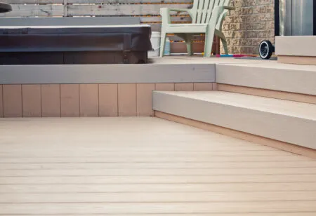 A wooden deck with steps, a brown hot tub, plastic chairs, and a brick wall in the background.