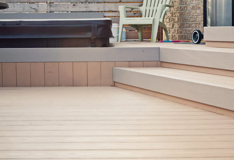 A wooden deck with steps, a brown hot tub, plastic chairs, and a brick wall in the background.